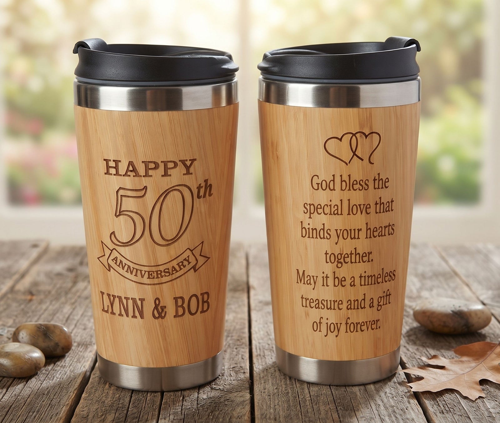 Two engraved stainless steel travel mugs with wood-look printed bodies are shown on a rustic wooden table. One mug is labeled 'HAPPY 50th ANNIVERSARY LYNN & BOB,' and the other has an engraved blessing with two hearts.