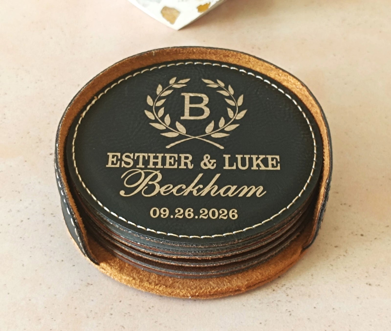 A stack of personalized leather coasters on a light beige surface. The top coaster is black with gold-embossed text reading 'ESTHER & LUKE Beckham 09.