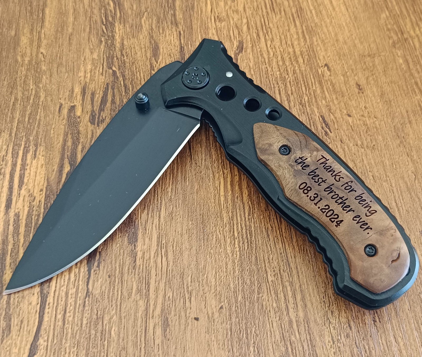 A folding pocket knife with a matte black blade and a black handle featuring a natural wood inlay engraved with 'Thanks for being the best brother ever. 08.