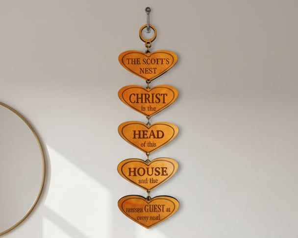 A vertical stack of five wooden heart-shaped wall plaques with religious and familial words ('CHRIST', 'HEAD', 'HOUSE', 'FAMILY FIRST') mounted on a light gray wall in a modern living room setting with a beige sofa and light wood flooring.