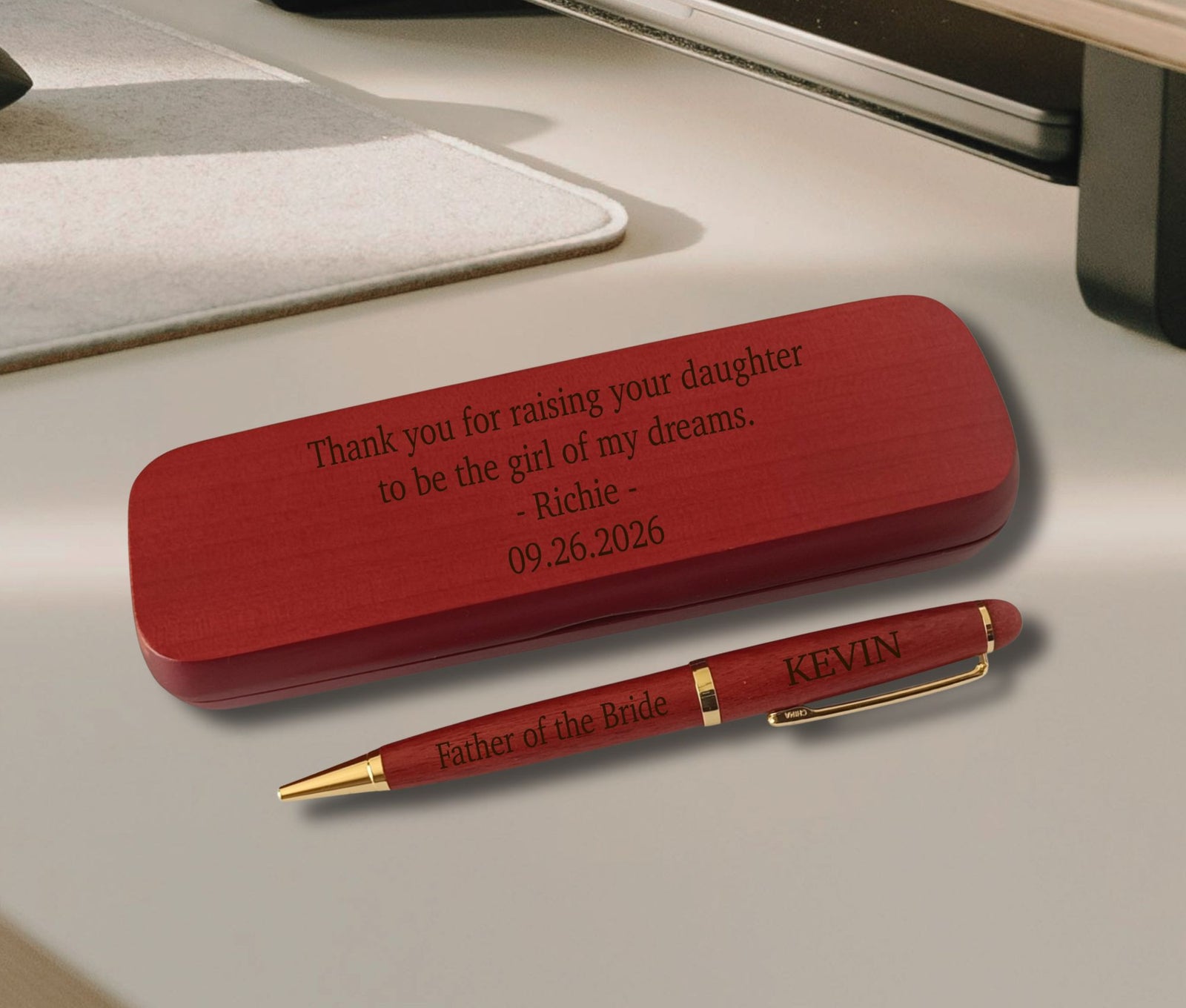 A personalized red wooden keepsake box and matching red pen with gold trim, placed on a light beige desk surface. The box is engraved with a thank-you message for raising a daughter, and the pen is engraved with 'Father of the Bride' and the name KEVIN.