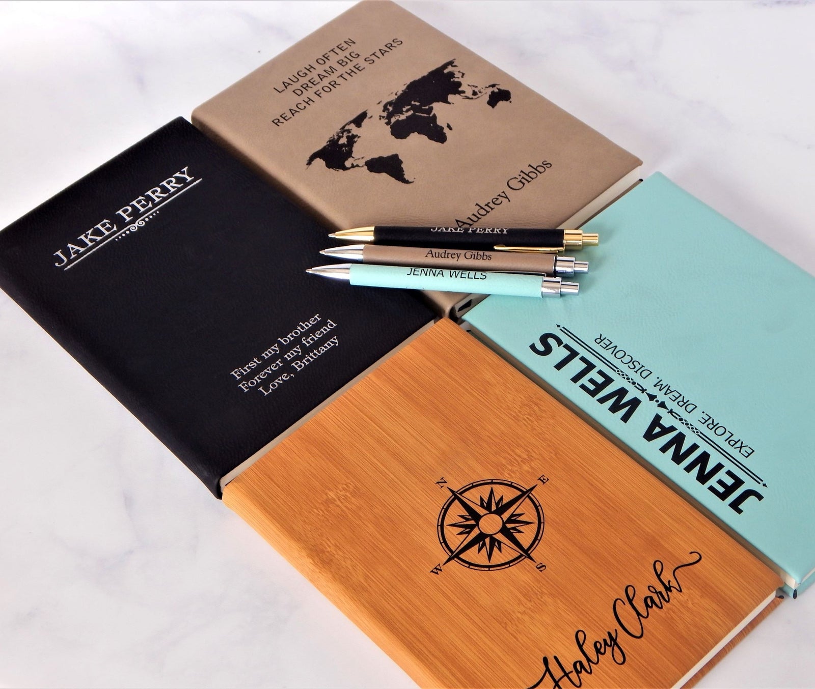 Four personalized hardcover notebooks in black, tan, light blue, and natural bamboo wood, each with a matching pen, arranged on a white marble surface. The notebooks feature custom names, travel-themed quotes, and black printed designs including a world map and compass rose.