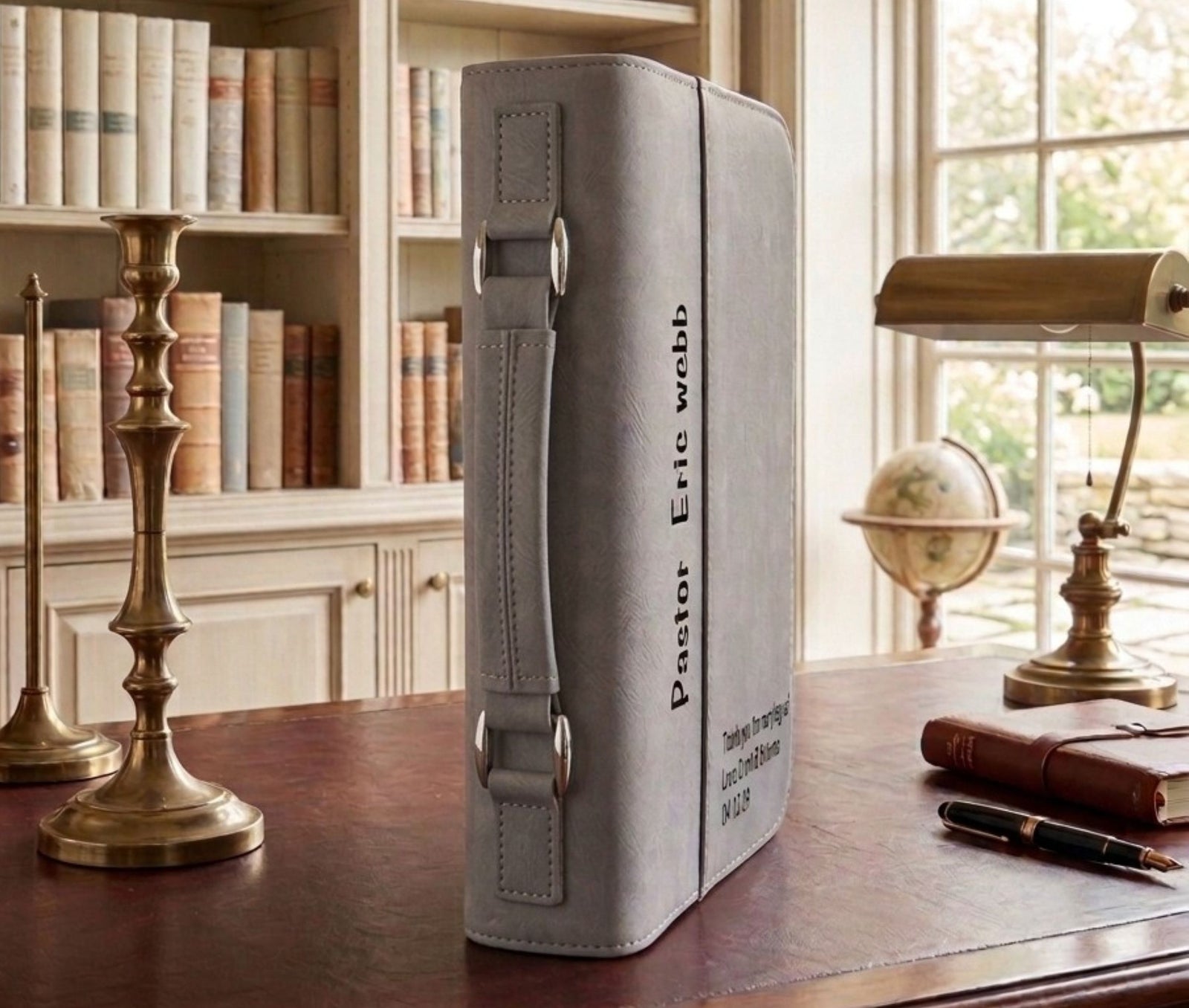 A gray faux-leather Bible cover standing upright on a wooden desk in a study setting, with bookshelves, brass candlesticks, a lamp, and a globe in the background. The cover features black printed text reading 'Pastor Eric wobb' and 'Believe the Word and Obey the Lord 2025'.