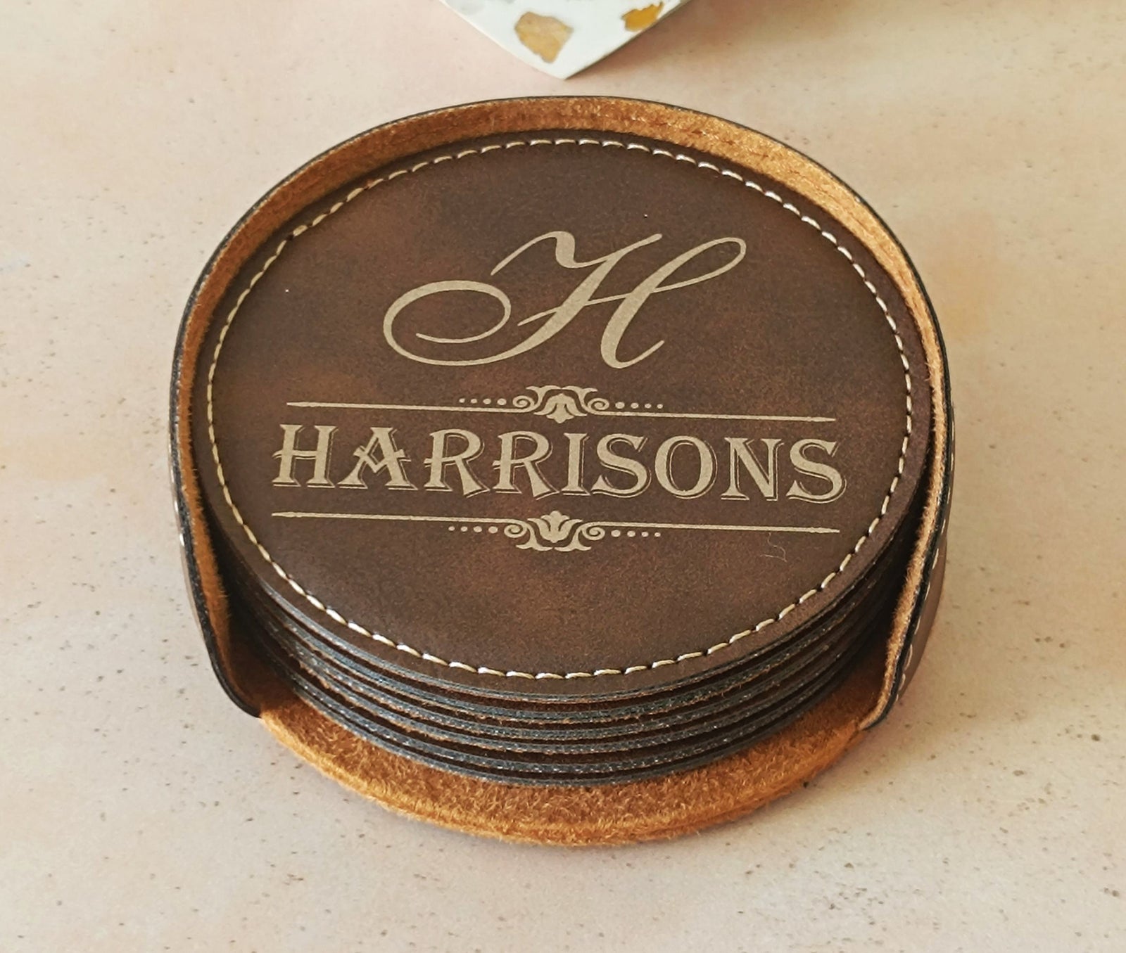 A stack of round brown leather coasters on a light beige surface. The top coaster is personalized with a cream-colored monogram 'H' and the name 'HARRISONS' in a serif font, surrounded by cream stitching.