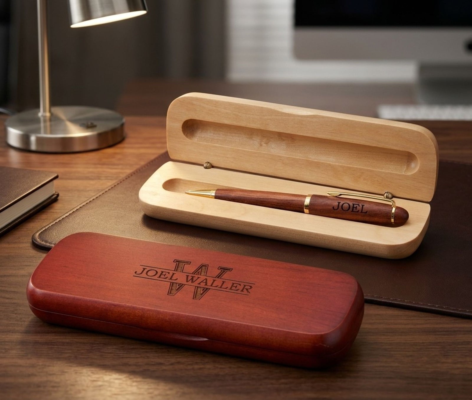 A personalized wooden pen set displayed on a desk, featuring an open light wood case holding a reddish-brown wooden pen engraved with 'JOEL', alongside a closed reddish-brown wooden case engraved with 'TWJ JOEL WALLER' on a dark wood desk.