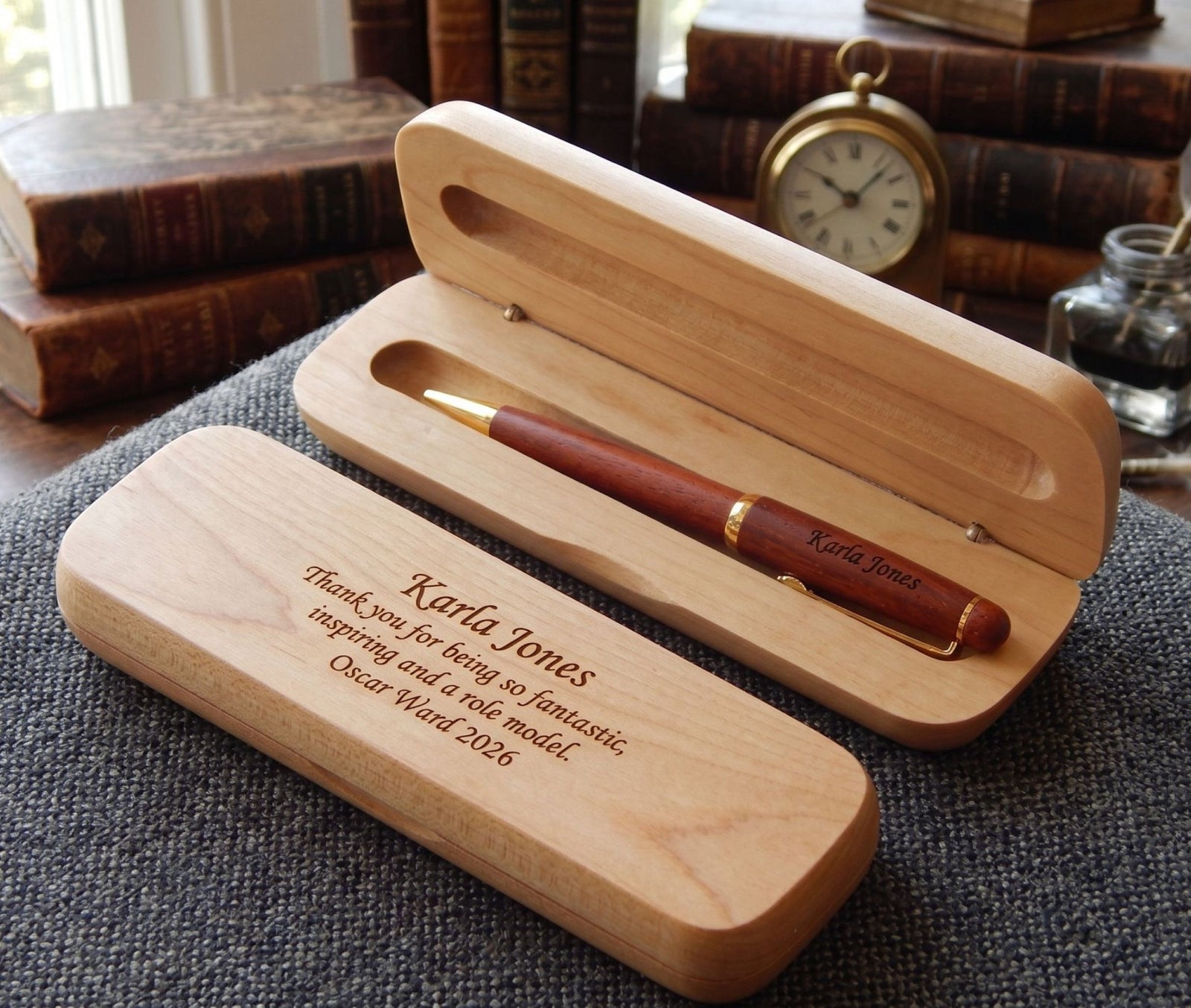 A personalized wooden pen gift set shown open and closed on a textured gray surface, with vintage books and a pocket watch blurred in the background. The set includes a light natural wood case engraved with the name 'Karla Jones' and a reddish-brown wooden pen with gold trim and 'Karla Jones' on the clip.