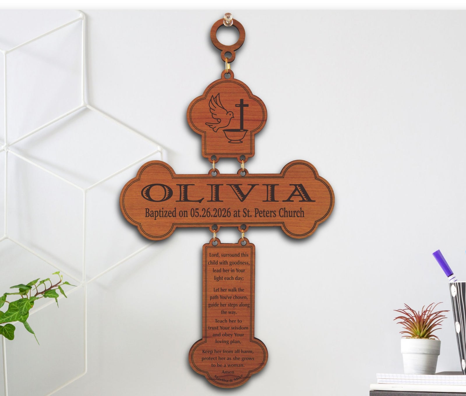 Personalized wooden cross wall plaque engraved with a dove and cross at the top, the name OLIVIA and baptism date at the center, and a scripture passage on a hanging lower section, shown mounted on a white wall with small plants and decor nearby.