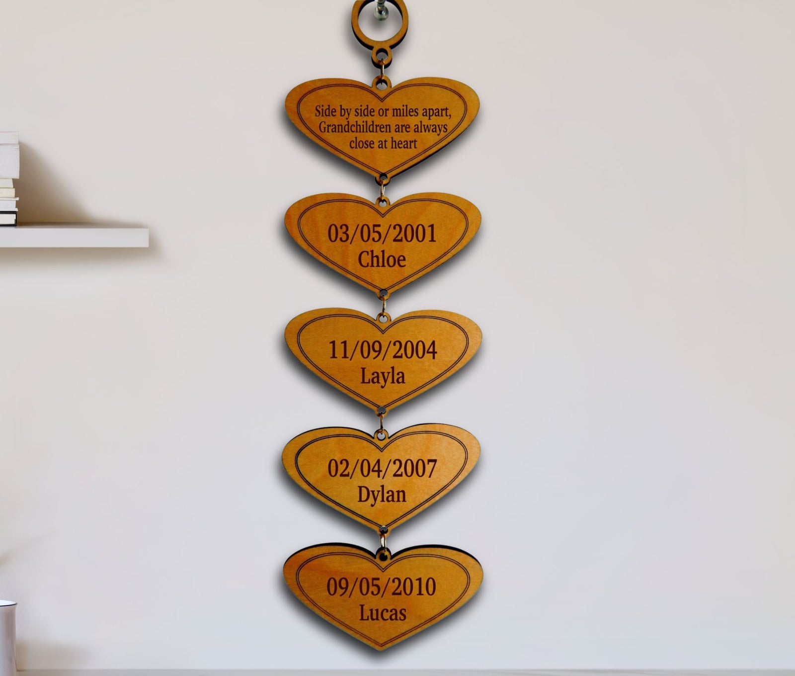 A vertical wooden hanging plaque made of five connected heart shapes, each engraved with a date and a child's name, displayed against a plain white wall.