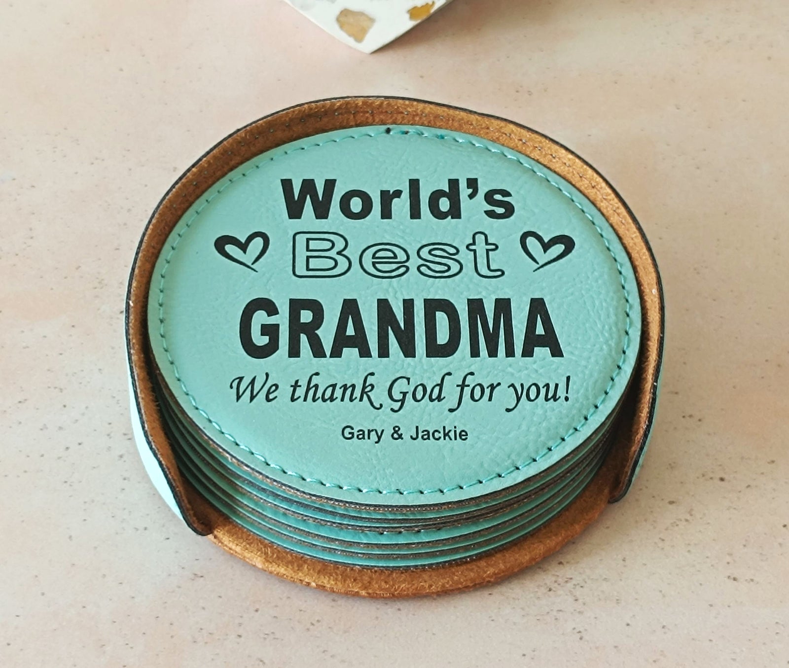 Personalized round engraved leather coasters displayed on a cream background. The top coaster is teal in color with white text reading 'World's best Grandma' ' we thank God for you and names below