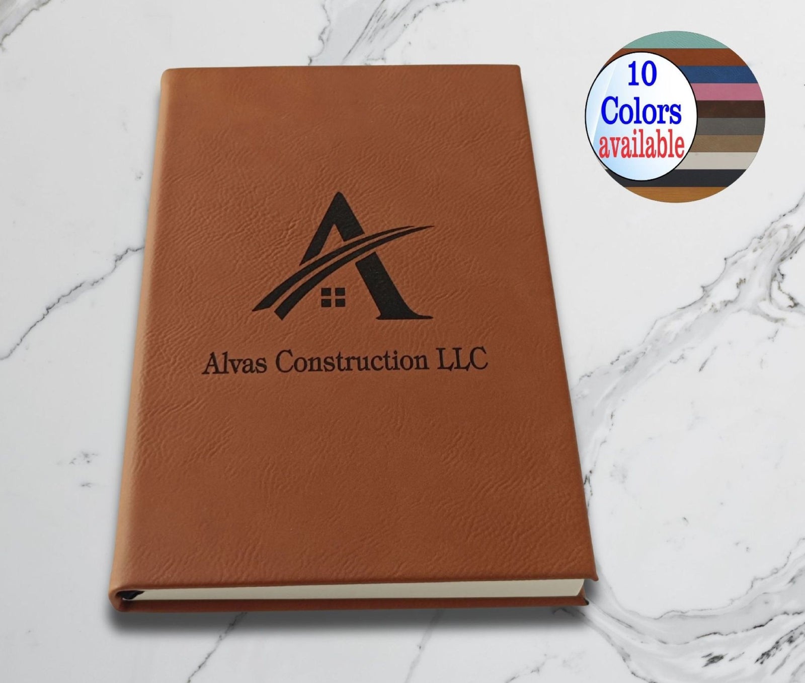A brown faux-leather hardcover notebook or journal with a black embossed logo and the text 'Alvas Construction LLC' on the cover, placed on a white marble surface. A circular badge in the top right says '10 Colors available'.