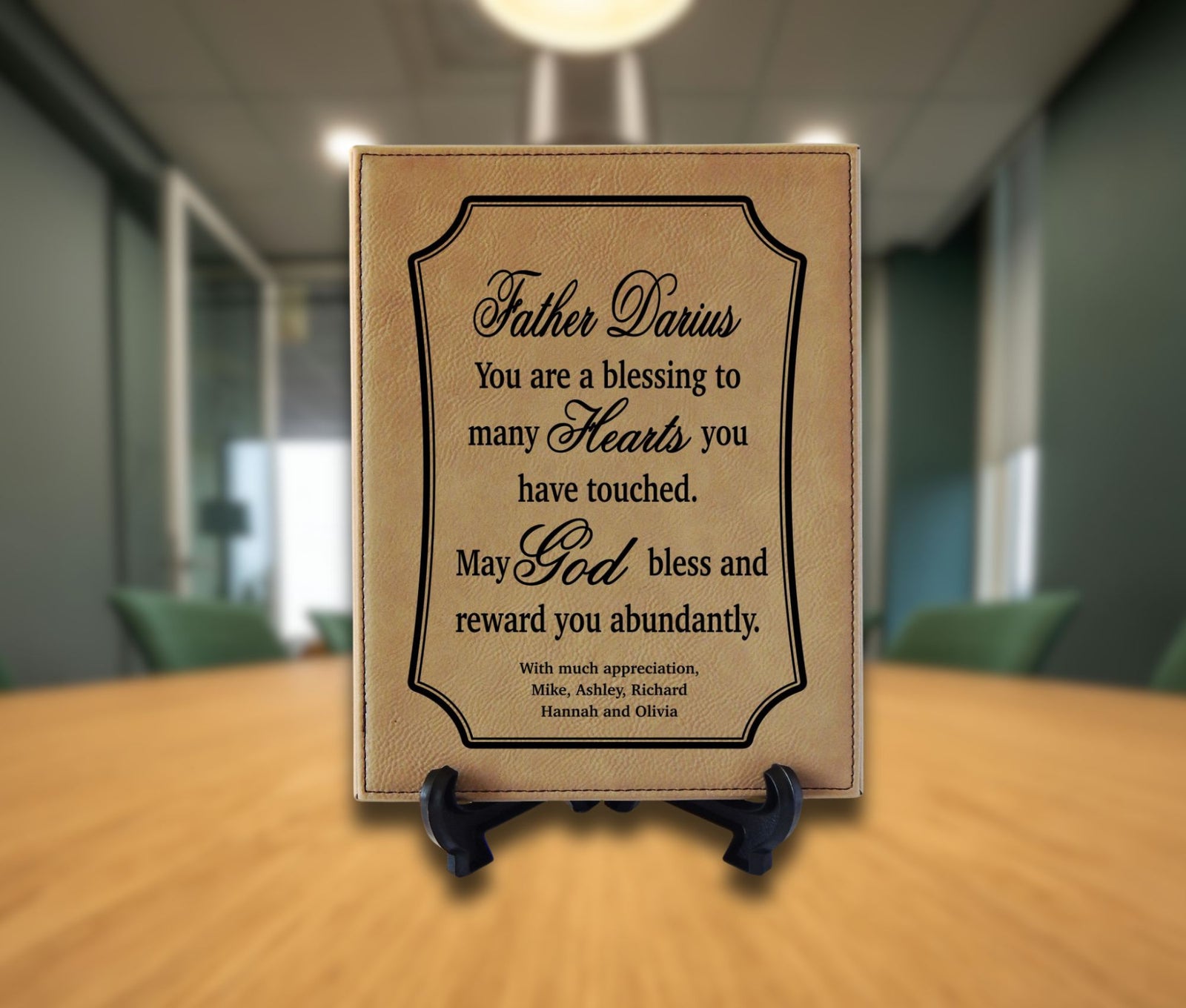 A personalized tan leather plaque with black text, displayed on a small black stand on a wooden table with a blurred office background.