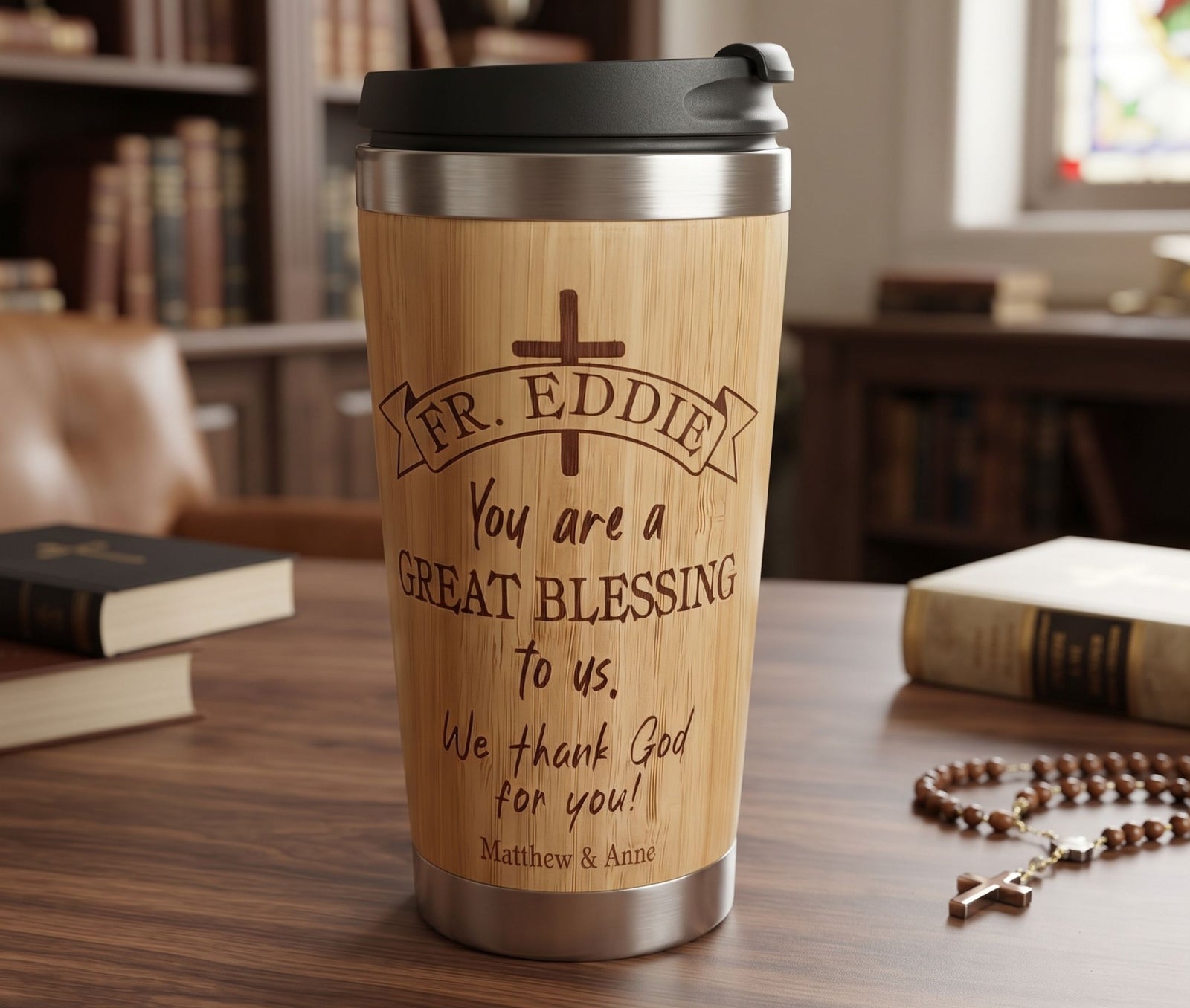 A bamboo travel tumbler with a black lid sits on a wooden table in a softly blurred library or study setting. The tumbler is engraved with a cross and the text 'FR.