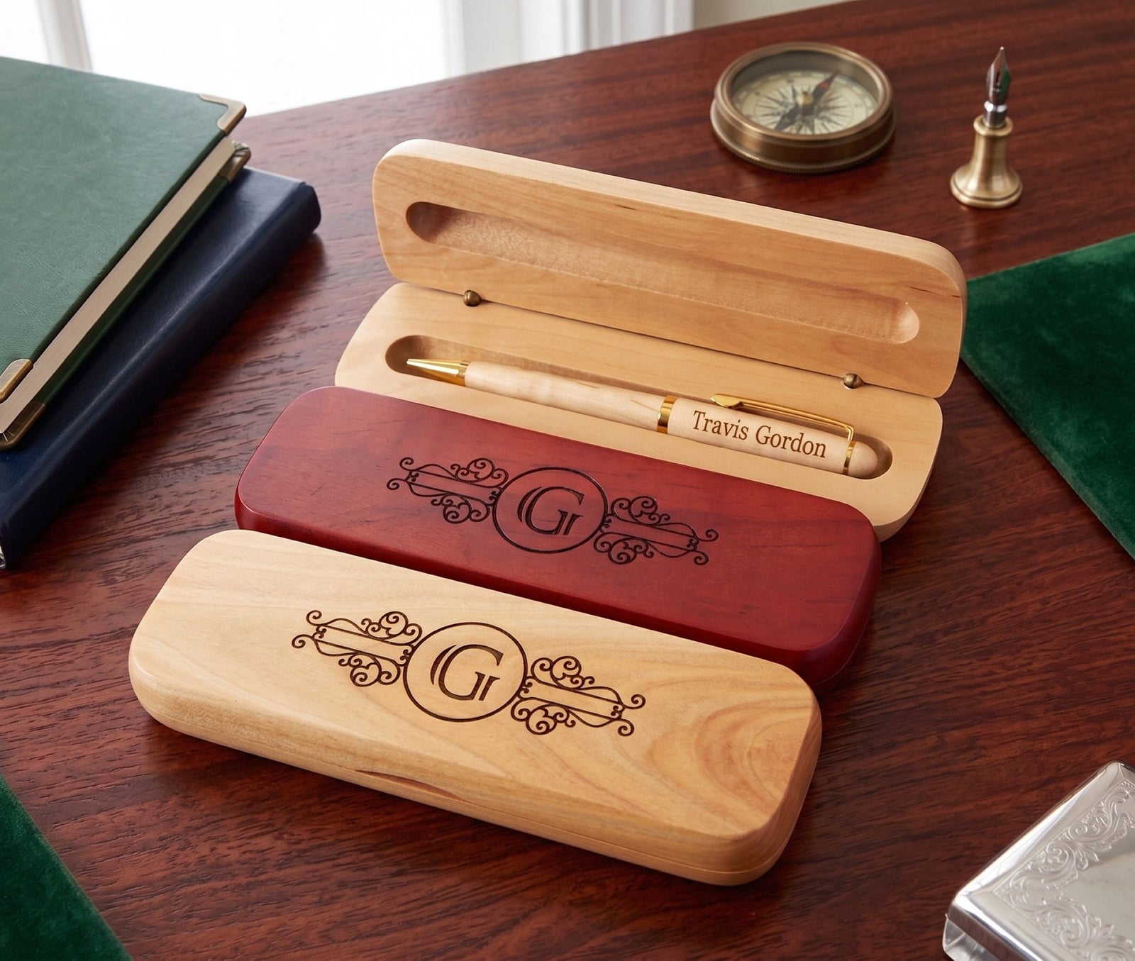 Three engraved wooden pen cases on a dark wood desk, with one open case holding a personalized wooden pen. The cases are shown in light natural wood, reddish wood, and a darker reddish-brown finish, each engraved with an ornate monogram and the text 'Travis Gordon'.