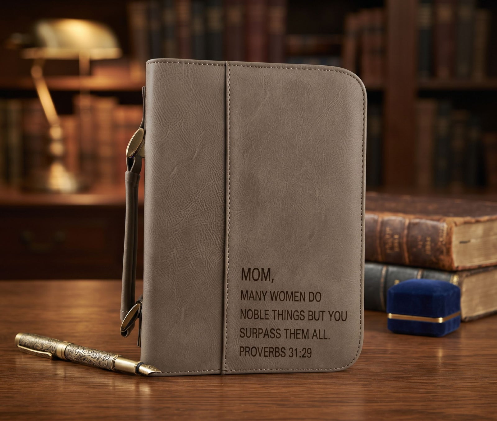 A gray faux-leather Bible cover or journal standing upright on a wooden table, with a pen resting in front and a blurred bookshelf background. The cover is engraved with the message: 'MOM, MANY WOMEN DO NOBLE THINGS BUT YOU SURPASS THEM ALL.