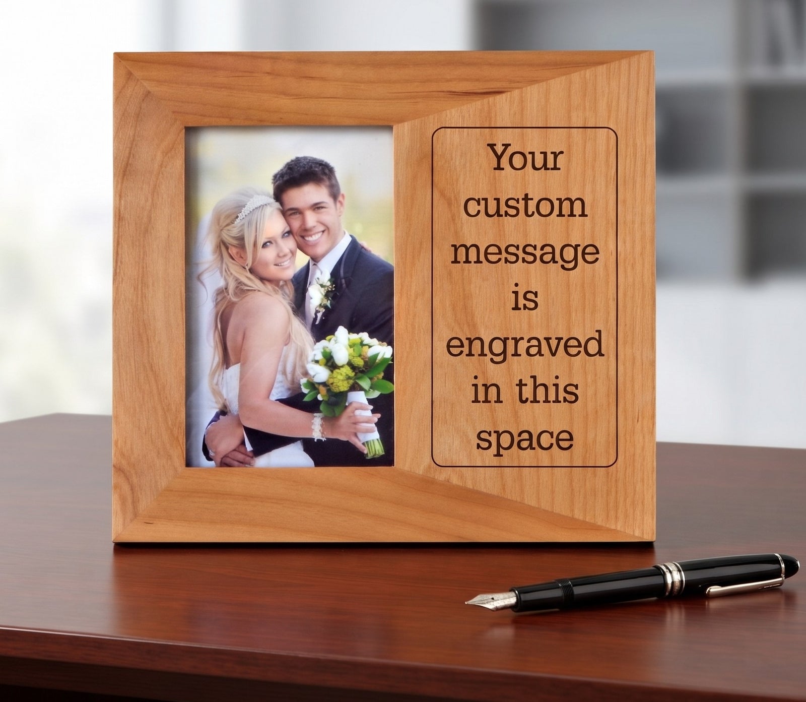 A wooden picture frame with a custom engraving slot, displayed on a dark wood table next to a black fountain pen. The photo insert shows a bride and groom, and the engraving on the right side reads 'Your custom message is engraved in this space'.