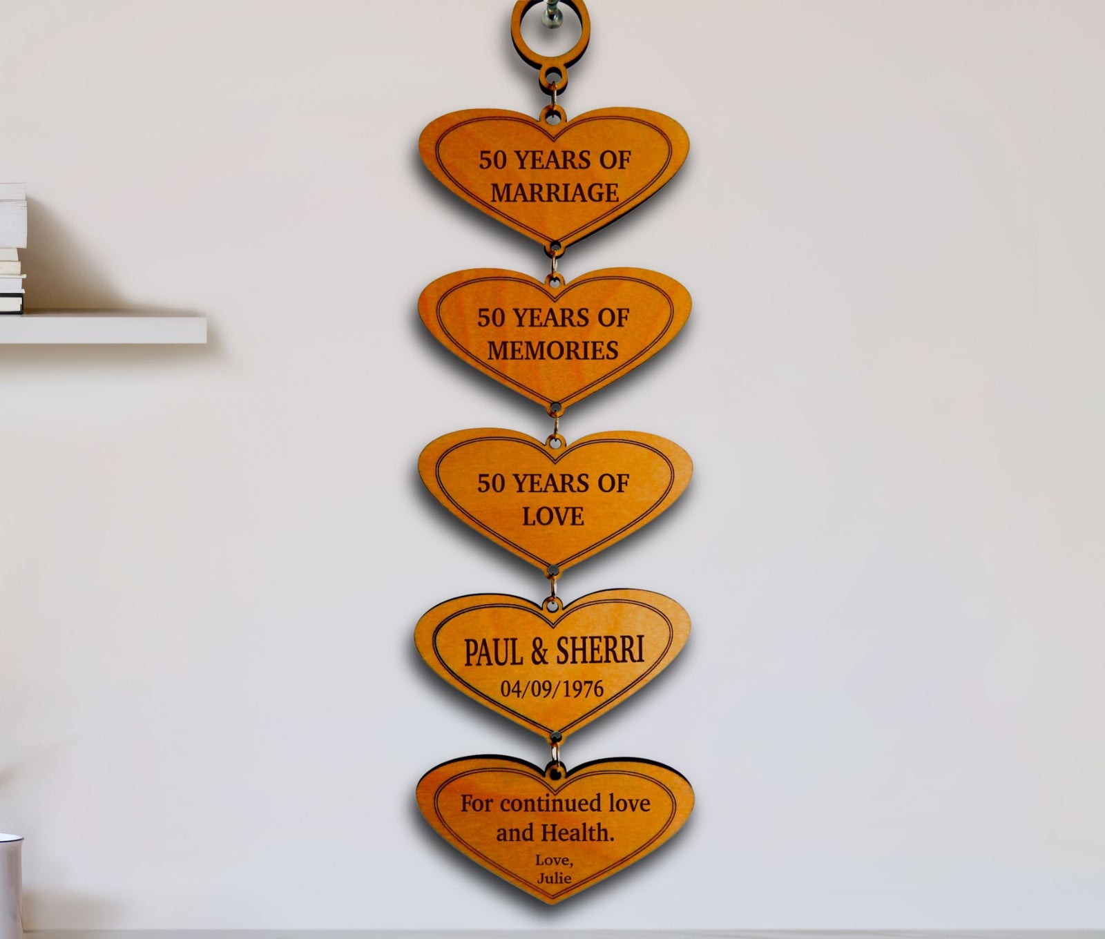 A vertical wooden heart-shaped wall hanging with five engraved gold-toned hearts stacked down, each reading a milestone phrase such as '50 Years of Marriage,' '50 Years of Memories,' '50 Years of Love,' 'Paul & Sherri 04/09/1976,' and 'For continued love and health.' The piece is mounted on a white wall.