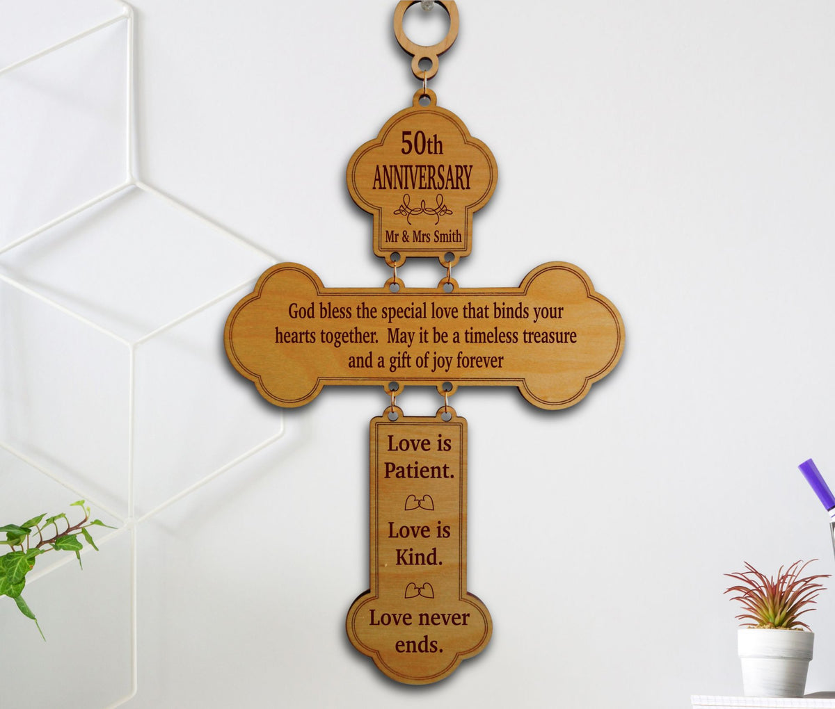 A wooden hanging cross-shaped wall plaque engraved with anniversary and quote text, shown hanging on a white wall with small plants nearby.