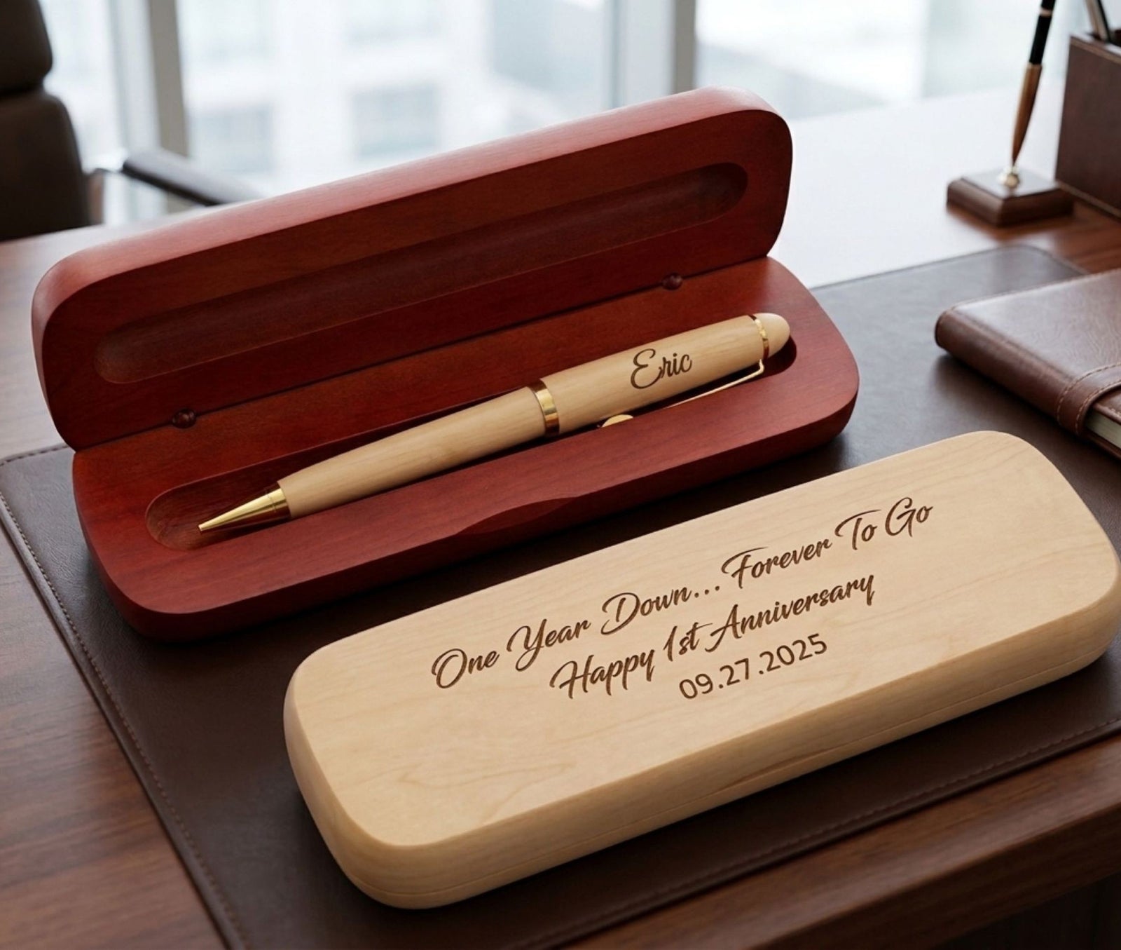 Personalized wooden pen gift set on a desk, featuring an open reddish-brown wooden case with a light wood pen inside, and a matching closed light wood case engraved with anniversary text in the foreground.