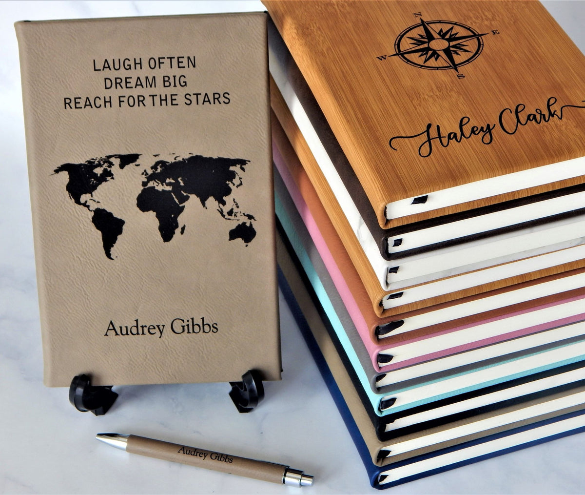 A stack of personalized hardcover notebooks and a matching pen on a light marble surface. The front notebook has a tan textured cover with black text reading 'LAUGH OFTEN DREAM BIG REACH FOR THE STARS' and a world map graphic, personalized with the name 'Audrey Gibbs.