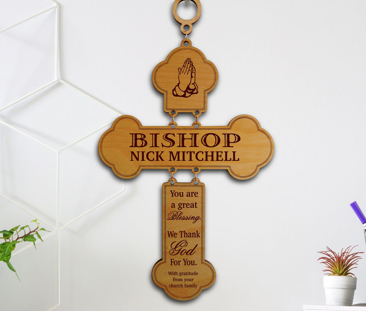 A personalized wooden cross wall hanging with a top loop, engraved with a praying hands icon and the text 'BISHOP NICK MITCHELL,' plus a hanging plaque that reads 'You are a great Blessing, We thank God For You. With gratitude from your church family.