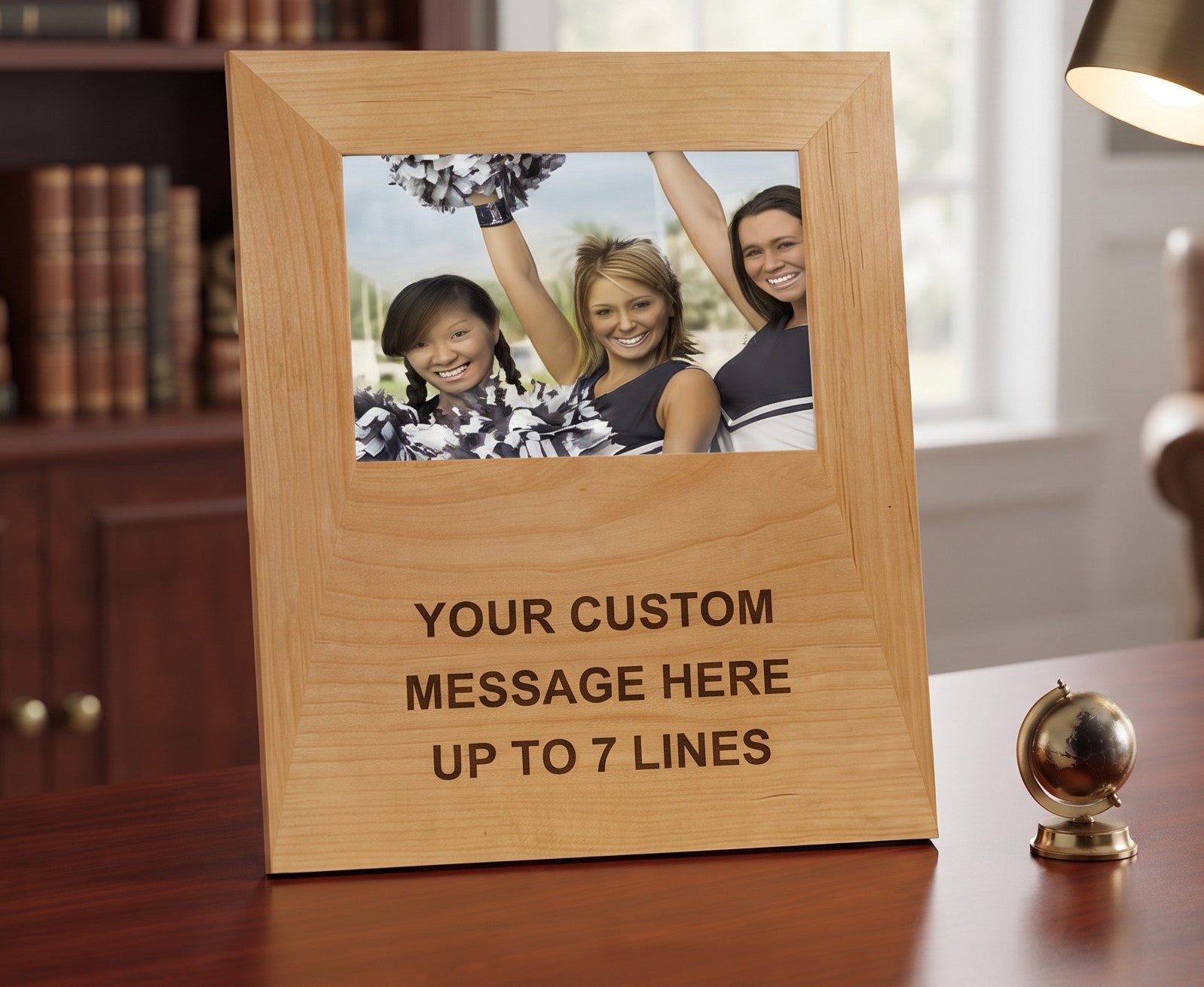 A personalized light wood picture frame standing on a wooden desk, with a sample photo of three cheerleaders at the top and engraved text reading 'YOUR CUSTOM MESSAGE HERE UP TO 7 LINES'. A bookshelf and lamp are visible in the background.