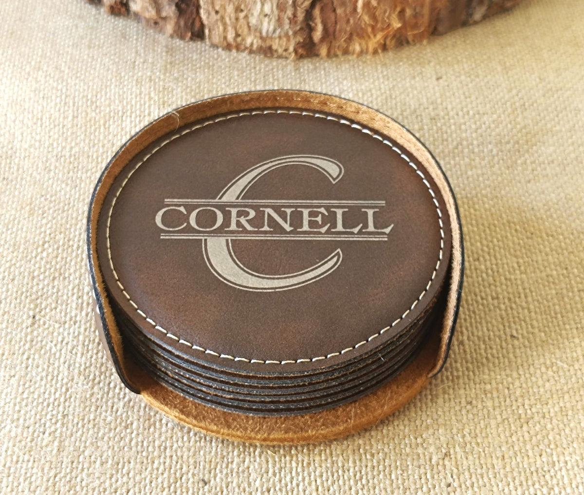 A stack of round brown leather coasters with white stitching, featuring a top coaster engraved with a large letter C and the word CORNELL, displayed on a beige fabric surface.