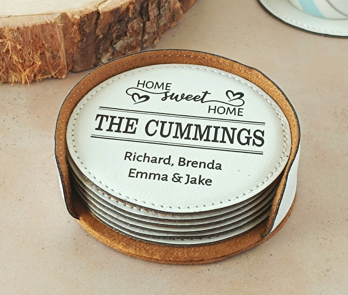 A stack of personalized round coasters with a white top coaster showing the text 'HOME sweet HOME THE CUMMINGS' and names, set on a beige surface with a rustic wood slice in the background.