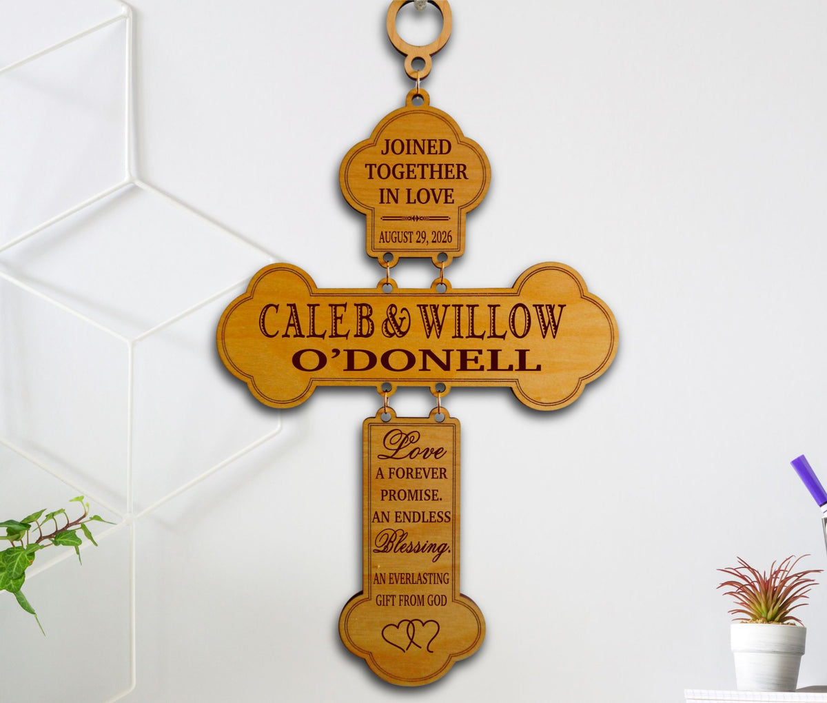 A personalized wooden cross wall hanging with engraved text reading 'CALEB &amp; WILLOW O'DONELL' and additional wedding-style inscriptions on the top and bottom sections, shown hanging against a white wall.
