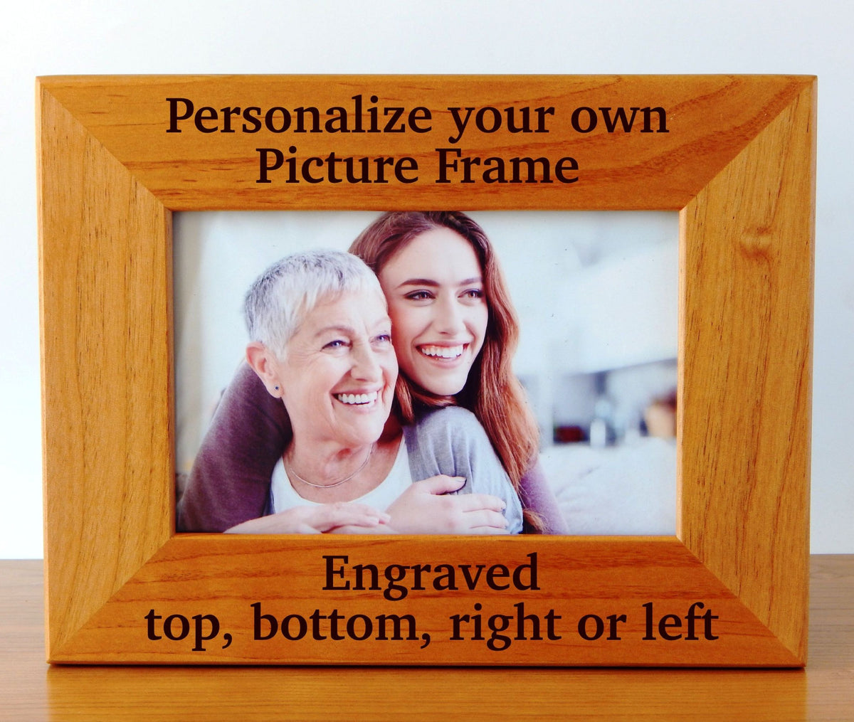A wooden picture frame with engraved text on the top and bottom reading 'Personalize your own Picture Frame' and 'Engraved top, bottom, right or left'. The frame is shown holding a photo of an older woman and a younger woman, set against a plain light background.