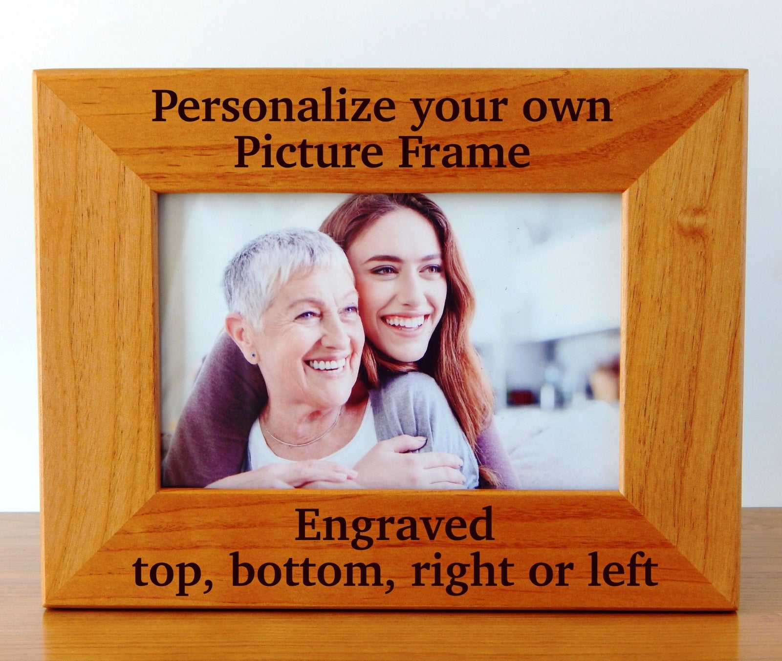 A wooden picture frame with engraved text on the top and bottom reading 'Personalize your own Picture Frame' and 'Engraved top, bottom, right or left'. The frame is shown holding a photo of an older woman and a younger woman, set against a plain light background.