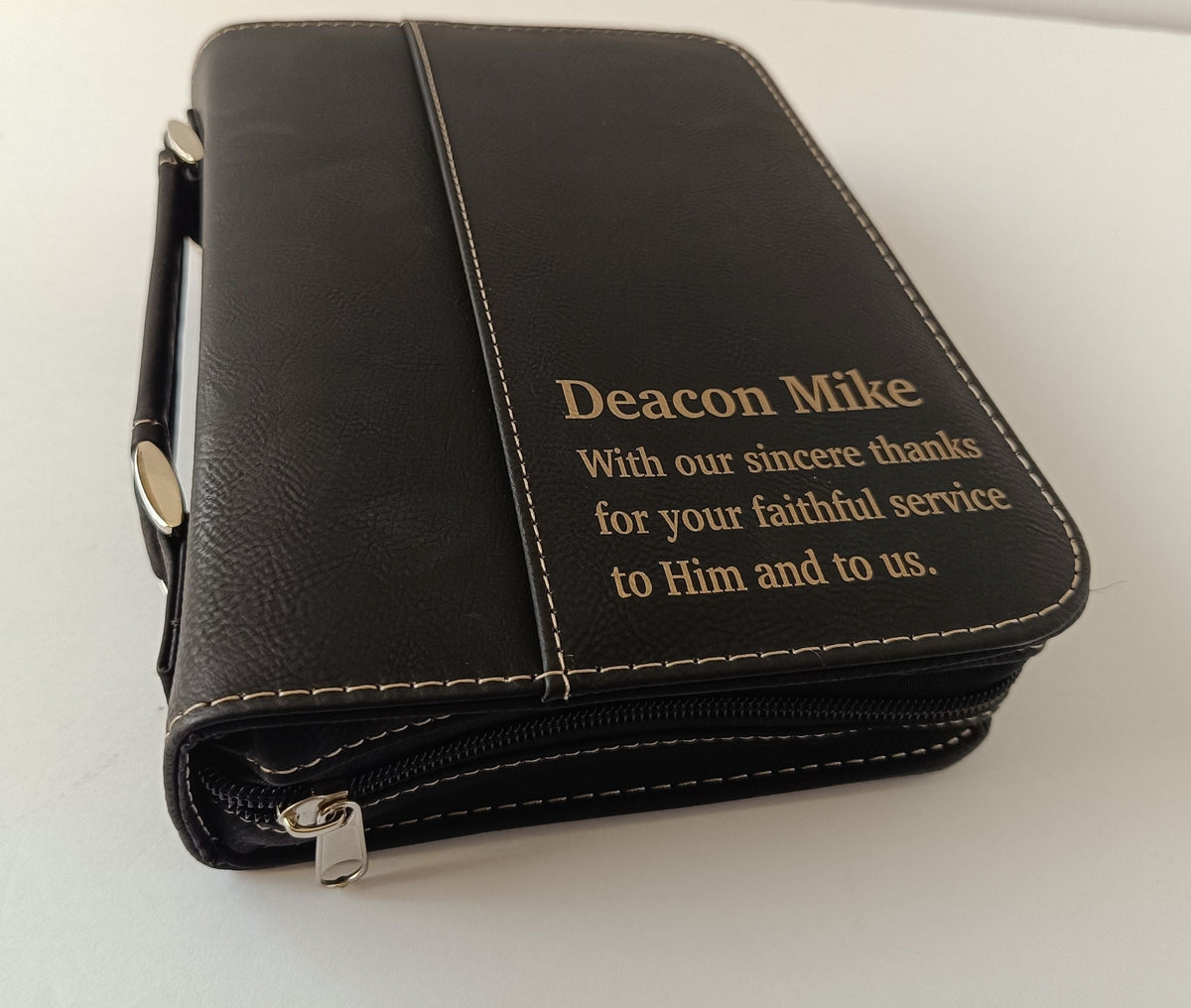 A black leather zippered Bible holder or wallet with gold embroidered text reading 'Deacon Mike With our sincere thanks for your faithful service to Him and to us.' on a plain white background.
