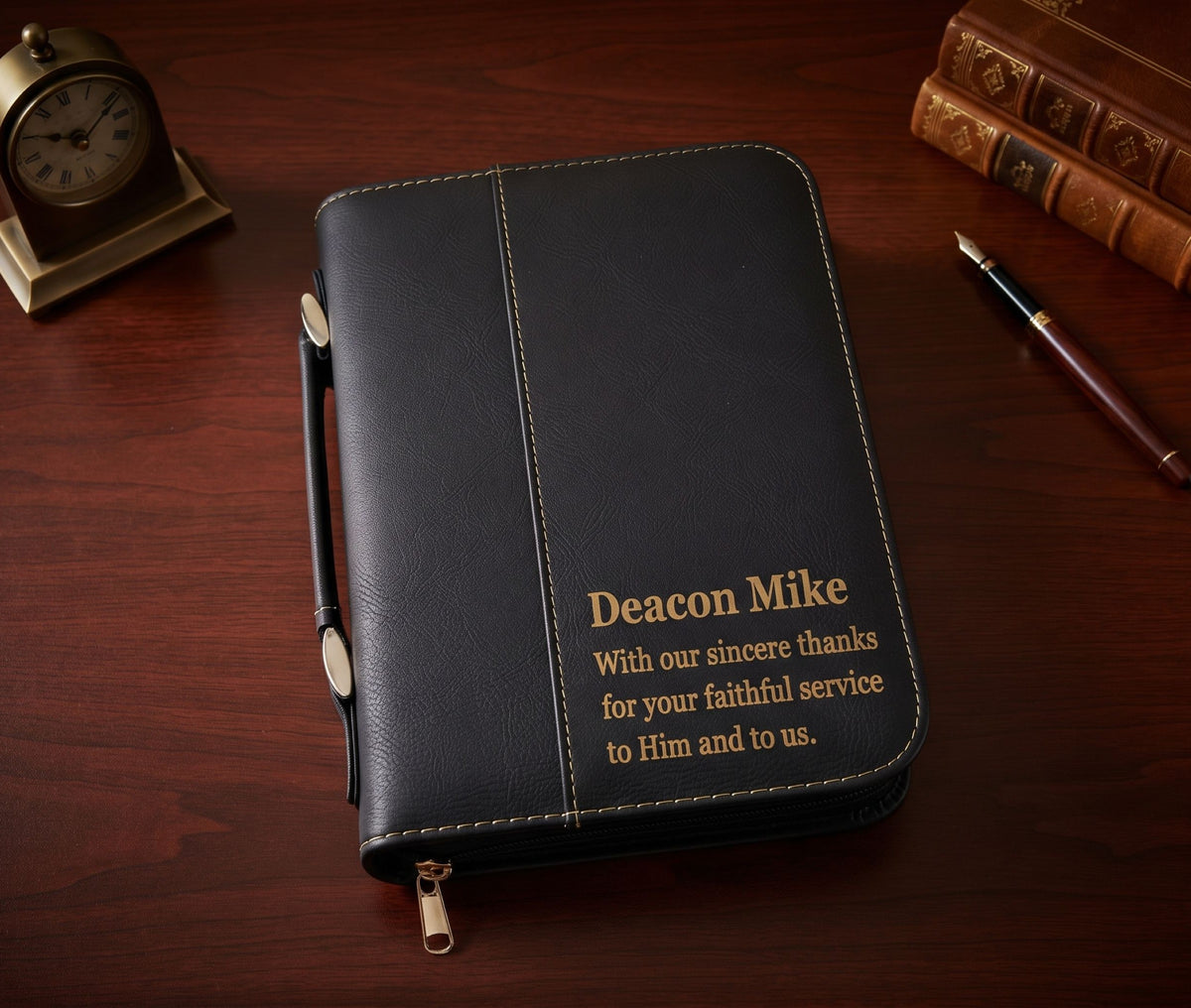 A black faux-leather zippered Bible or prayer journal with gold text reading 'Deacon Mike' and a thank you message, placed on a dark wooden desk with a pen, books, and a clock nearby.