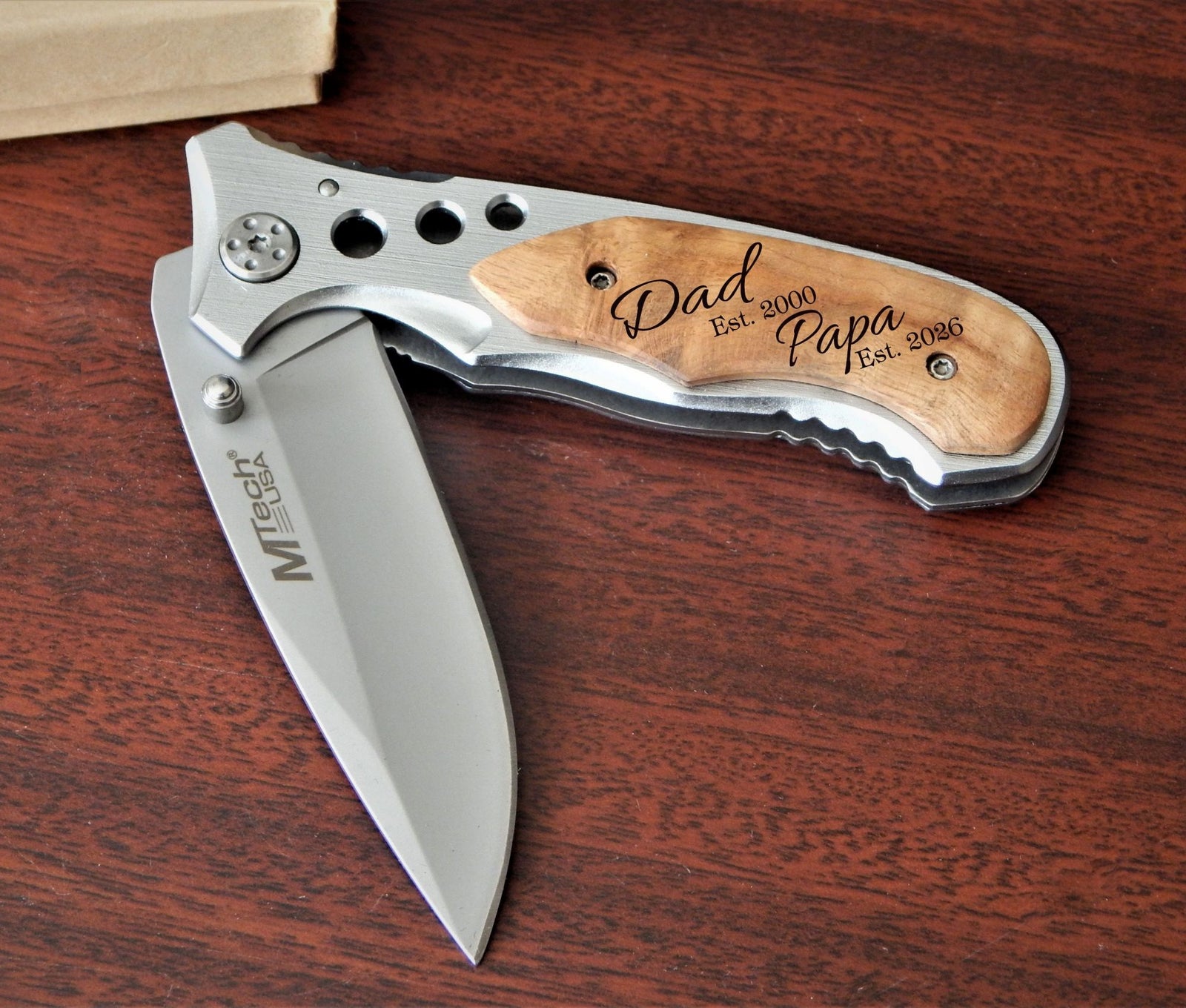 A folding pocket knife with a silver stainless steel blade and handle, featuring a light wood inlay engraved with 'Dad Papa Est. 2000'.