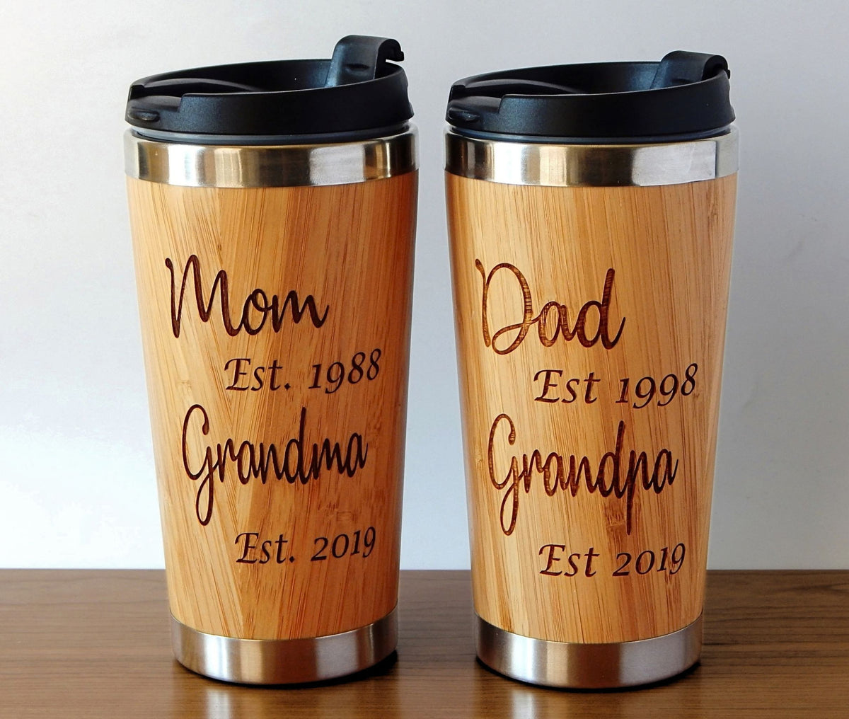 Two personalized bamboo travel mugs with black lids, displayed side-by-side on a wooden table against a neutral light gray background. One mug is engraved with 'Mom Est.