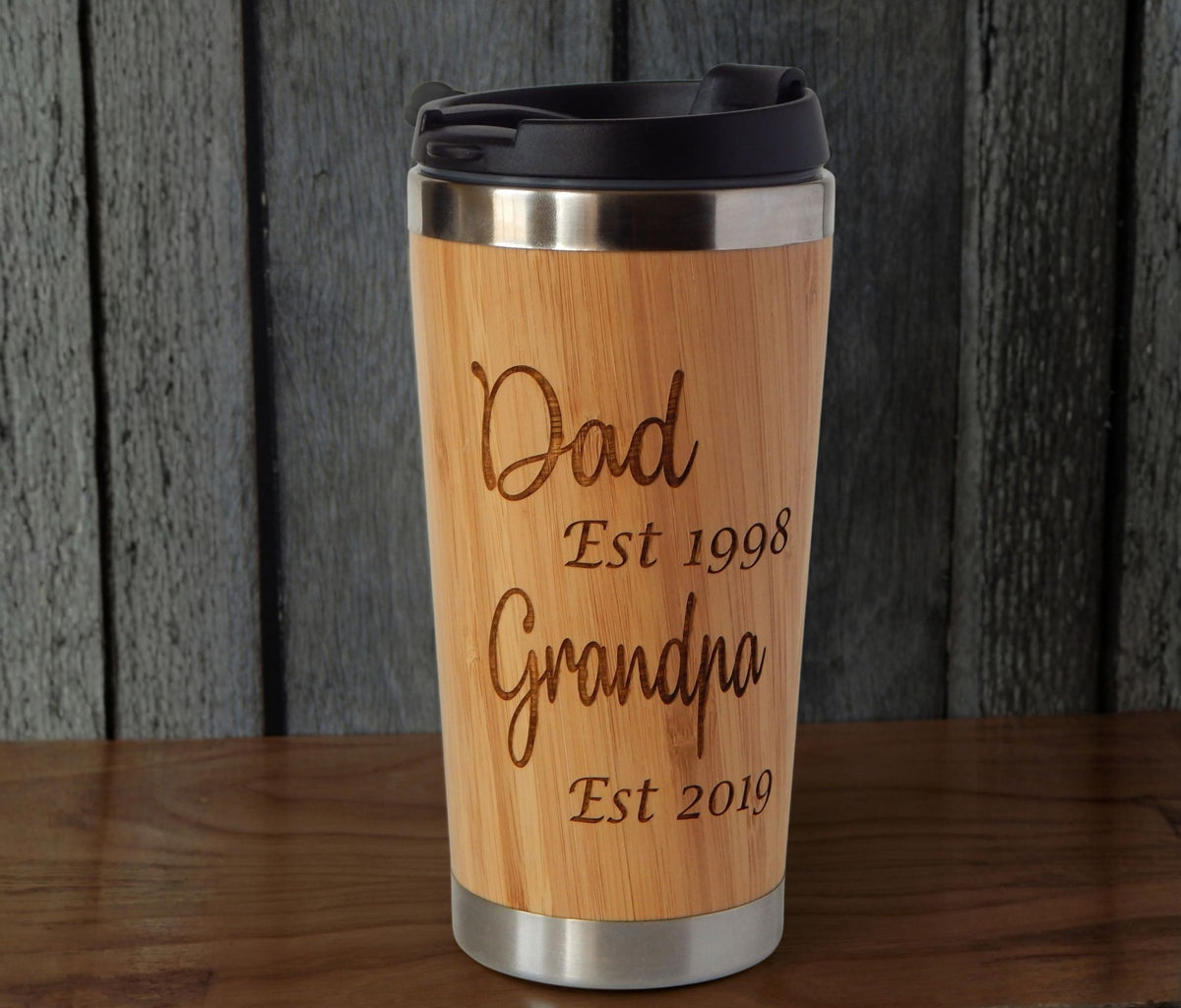 A personalized bamboo travel tumbler with a black lid, featuring engraved text reading 'Dad Est 1998 Grandpa Est 2019', set against a dark wood plank background.