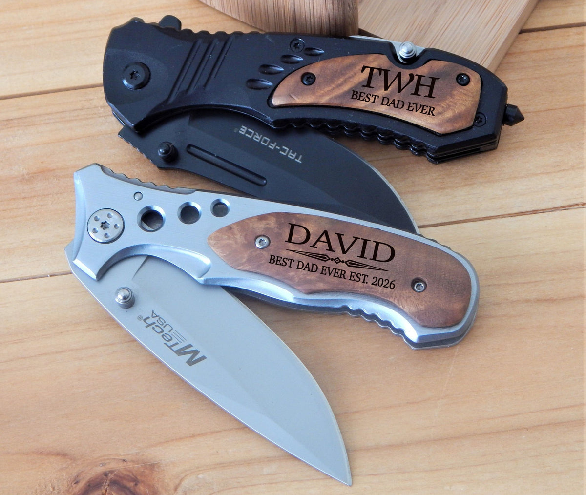 Two folding pocket knives displayed on a light wood surface. The top knife is black with a brown wood inlay engraved with 'TWH BEST DAD EVER'.