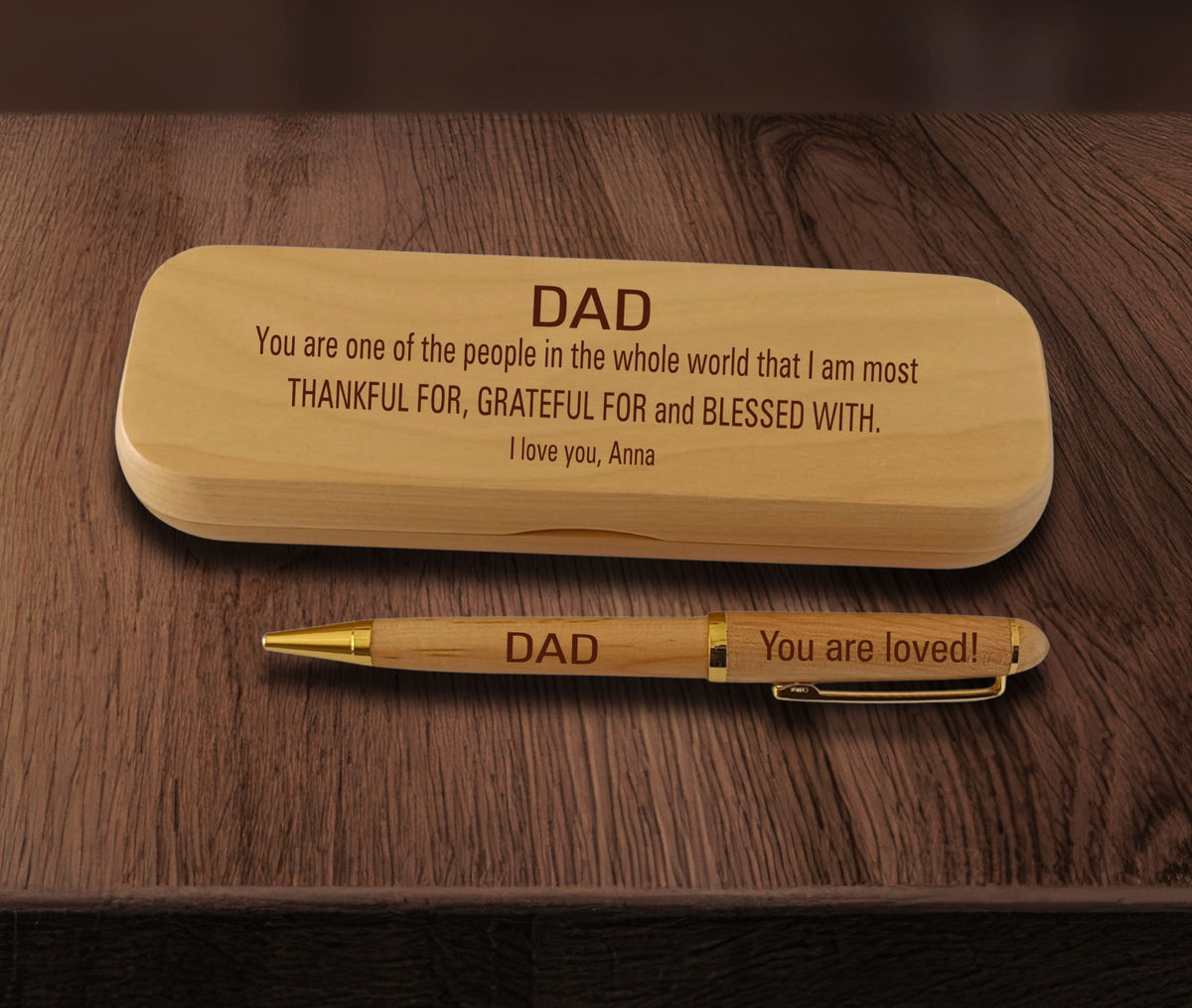 A personalized wooden pen engraved with 'DAD' and 'You are loved!' sits on a dark wood table beneath a matching engraved wooden box.