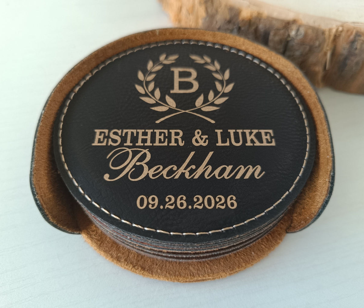A personalized leather coaster with a round black top featuring gold monogrammed text and a laurel wreath, resting on a stack of matching brown leather coasters on a light surface.
