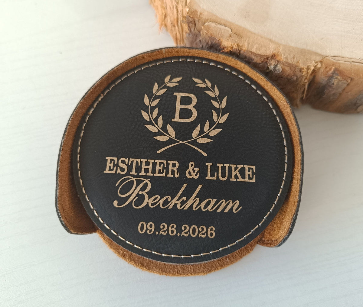A round black leather coaster with personalized gold-toned engraving, featuring a laurel wreath with the letter B, the names Esther &amp; Luke Beckham, and the date 09.26.