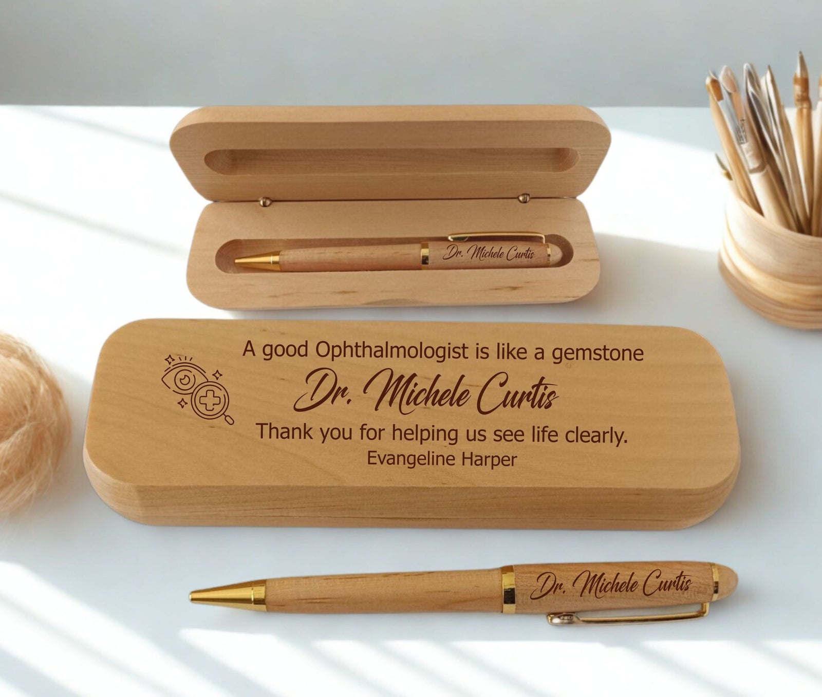 Personalized wooden pen gift set with engraved text for an ophthalmologist, shown with an open wooden case, a closed engraved case, and the pen on a white surface with soft natural shadows.