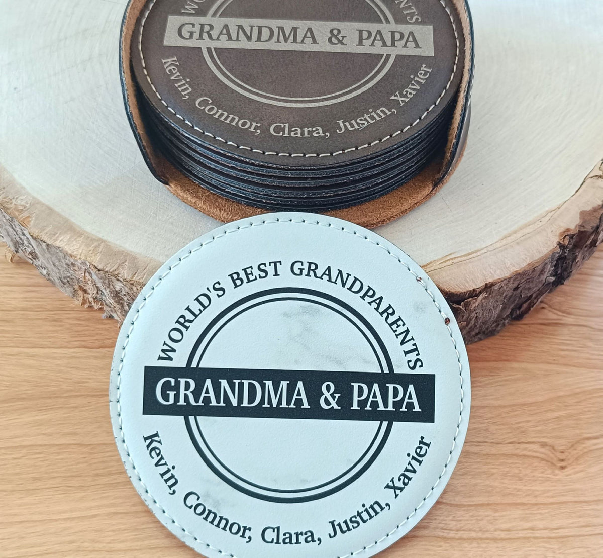 Two personalized round coasters displayed on a wood slice background. The top coaster is dark brown with white text reading 'GRANDMA &amp; PAPA' and names around the edge.
