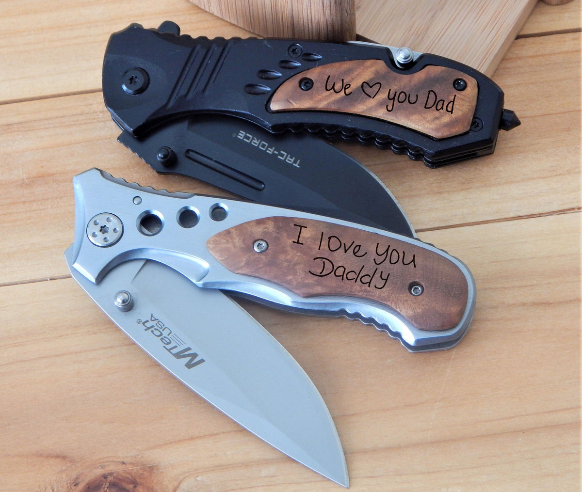 Personalized folding pocket knives handwriting gift grandpa or dad. Displayed on a light wood surface. The top knife is black with a brown wood handle inlay reading 'We love you Dad'.