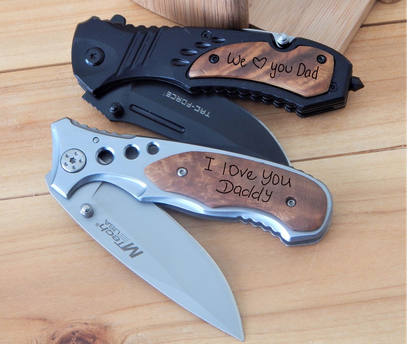 Personalized folding pocket knives handwriting gift grandpa or dad. Displayed on a light wood surface. The top knife is black with a brown wood handle inlay reading 'We love you Dad'.