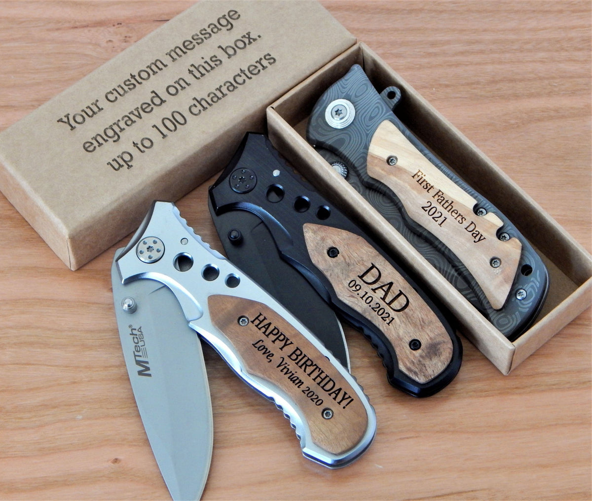 Three personalized folding pocket knives displayed on a wooden surface next to their kraft gift box. One knife is silver with a wood inlay engraved 'HAPPY BIRTHDAY! Love Trevor 2021', one is black with a wood inlay engraved 'DAD 09.