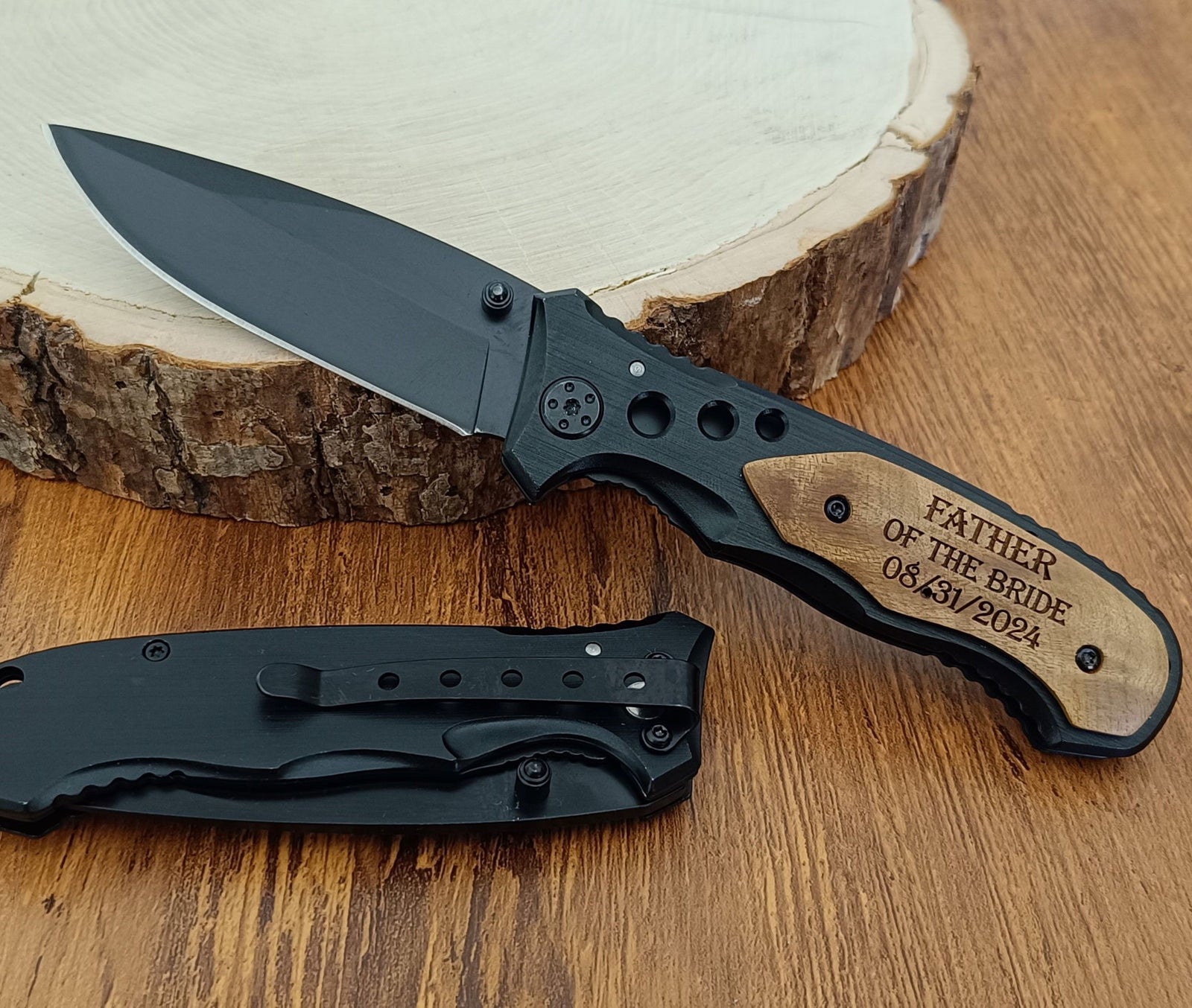 A black folding pocket knife with a wood inlay handle is displayed open on a wooden surface next to its black sheath. The handle features a custom engraved wood inlay with the text 'FATHER OF THE BRIDE 05.