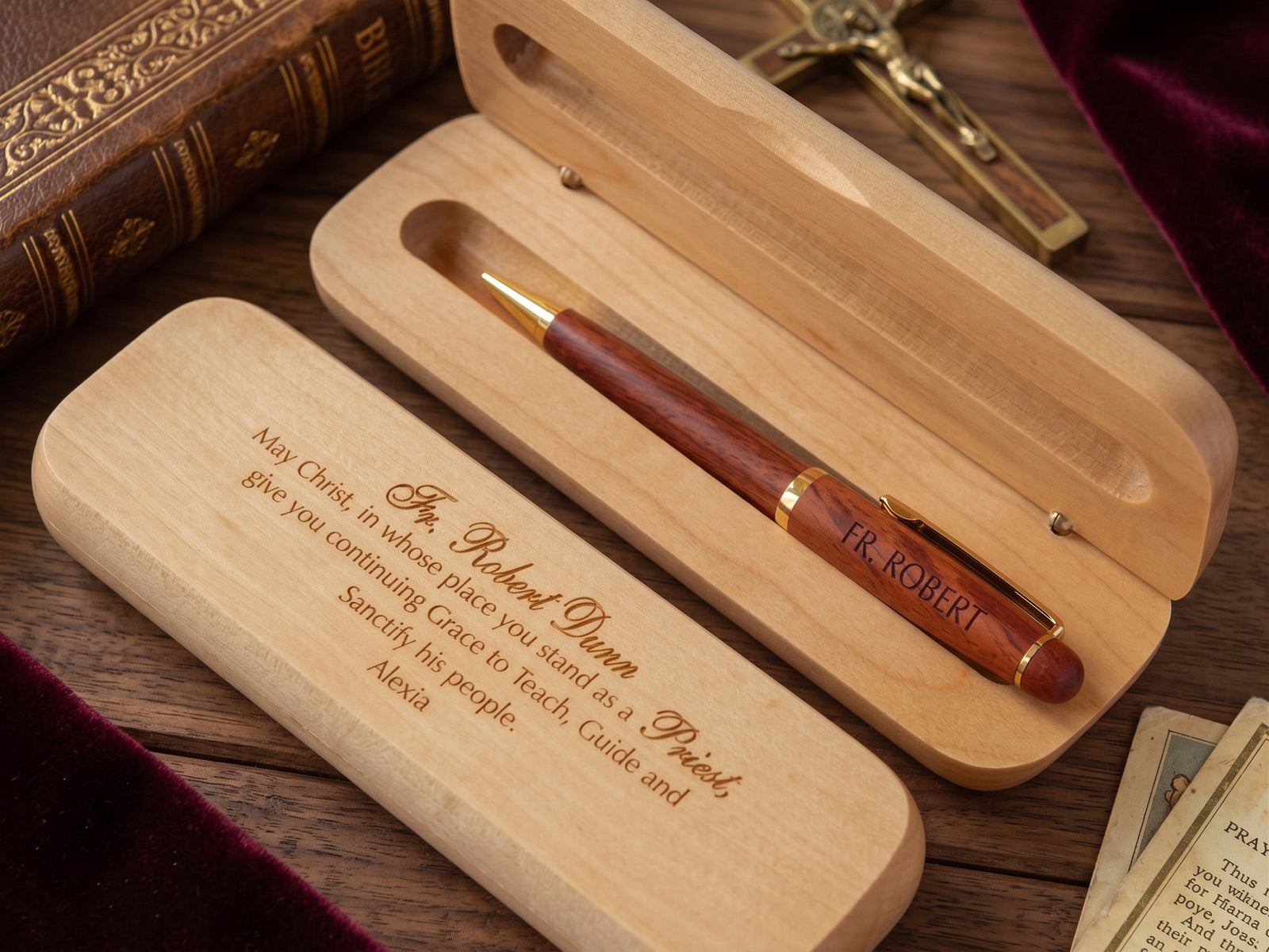 A personalized wooden pen with a reddish-brown wood body and gold trim, presented in an open light wood box engraved with a prayer. The box is placed on a wooden table with a leather-bound book, a crucifix, and cards nearby.