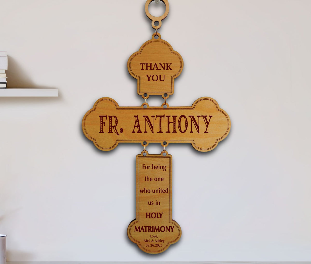 A wooden hanging cross plaque engraved with 'THANK YOU FR. ANTHONY' and a Holy Matrimony message, displayed against a white wall.