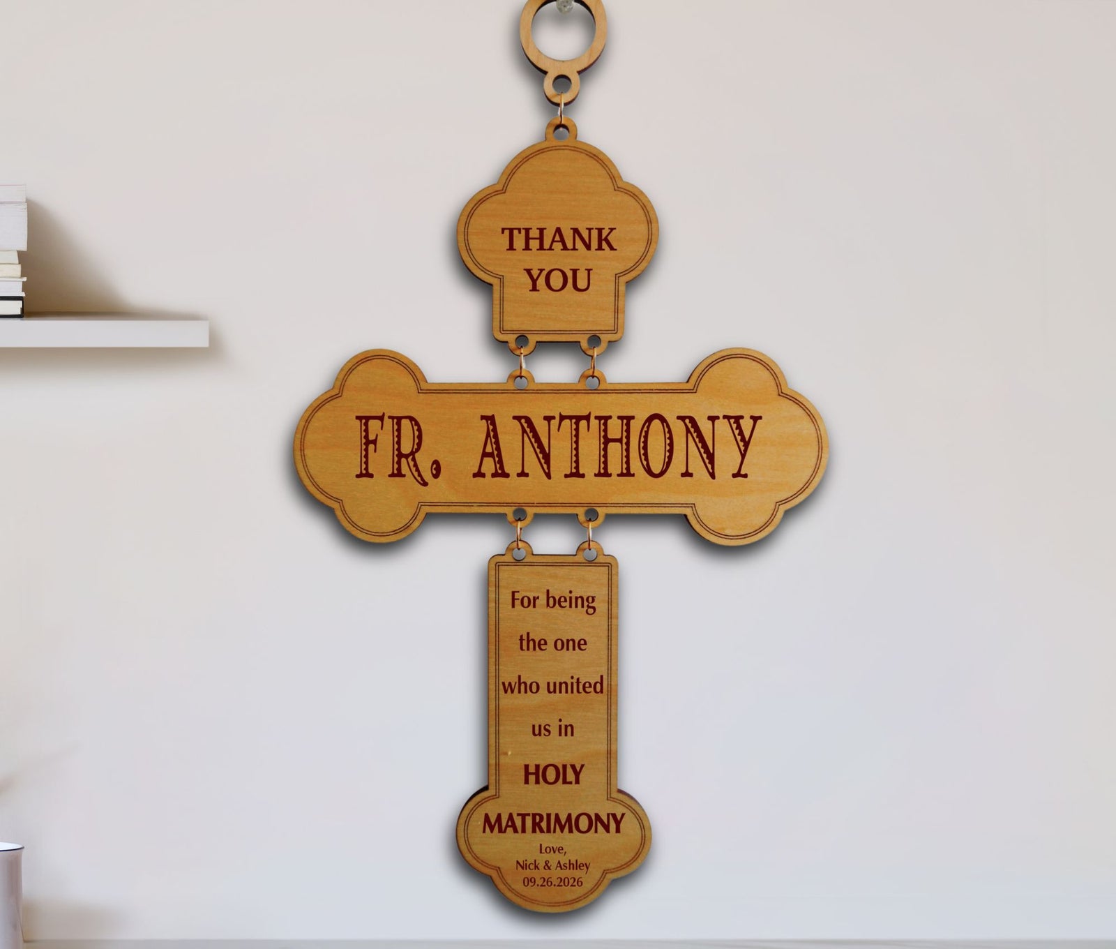 A wooden hanging cross plaque engraved with 'THANK YOU FR. ANTHONY' and a Holy Matrimony message, displayed against a white wall.