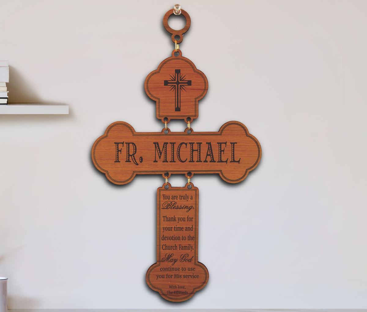A personalized wooden cross wall hanging with a top loop, engraved cross, the name 'FR. MICHAEL' across the center, and a hanging plaque with a blessing message, shown mounted on a light wall.