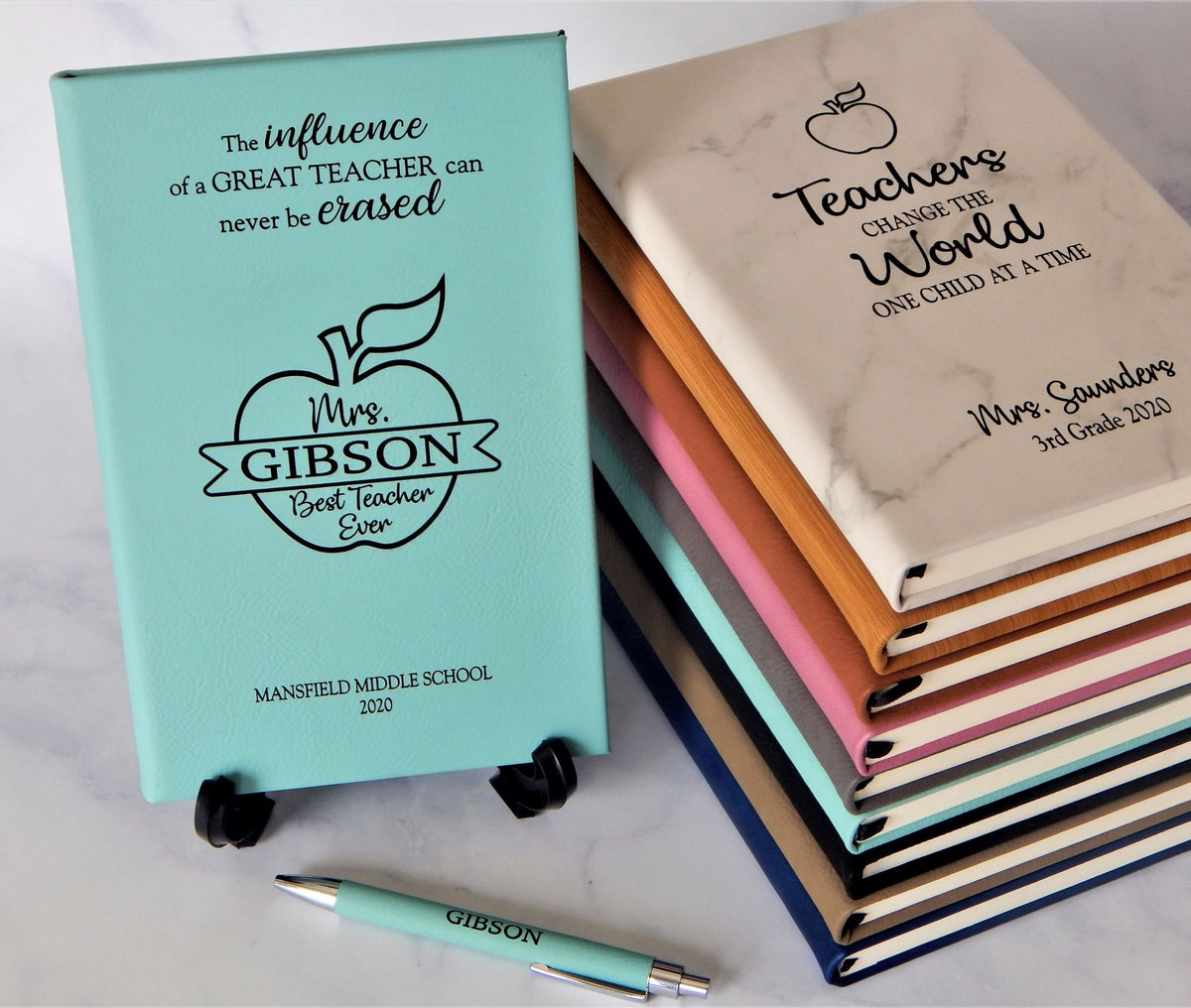 Personalized hardcover notebooks for teachers displayed on a light marble surface, with one mint blue notebook standing upright and a stack of colorful notebooks fanned out behind it. A matching mint blue pen lies in front.